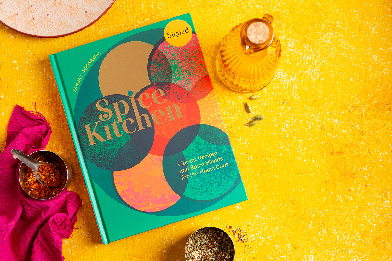 Spice Kitchen cookbook by Sanjay Aggarwal, on a yellow background with spices and a pink cloth.