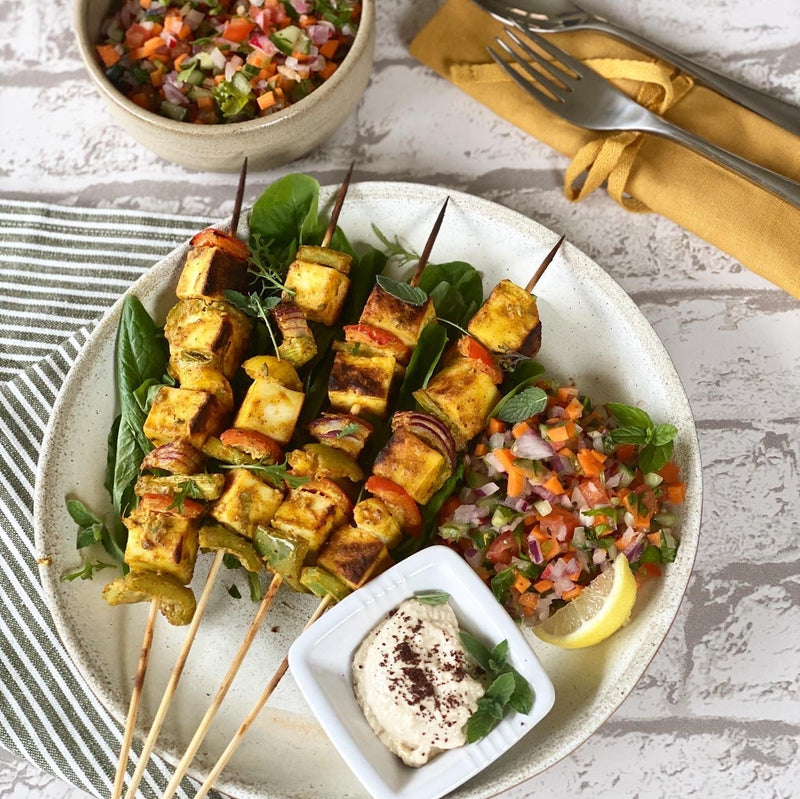 Delicious paneer tikka served on bamboo skewers with a fresh yoghurt side dish, tangy salsa, baby leaves and a slice of lemon