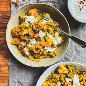 A bowl of cauliflower and potato curry on a grey tea towel with a spoon and glass of water