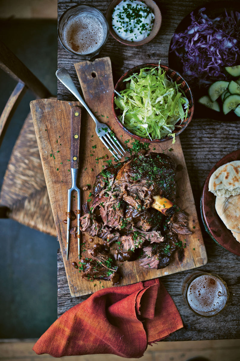 A gorgeous Tender partly shredded lamb joint on a chopping board surrounded by a Pita, Salad and dips. 