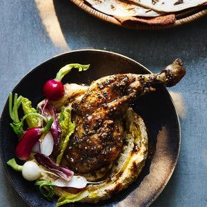 Roasted chicken leg with Za'atar spice blend and salad on a bed of hummus. There's a bowl of pitta breads. All shot on a blue background