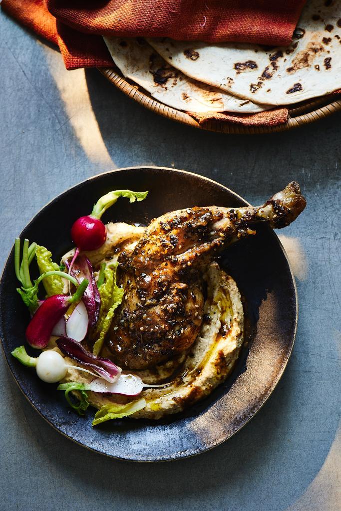 Roasted chicken leg with Za'atar spice blend and salad on a bed of hummus. There's a bowl of pitta breads. All shot on a blue background