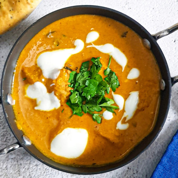 A pan of Butter Chicken Curry garnished with dollops of greek yoghurt and fresh coriander, with a naan bread on the side