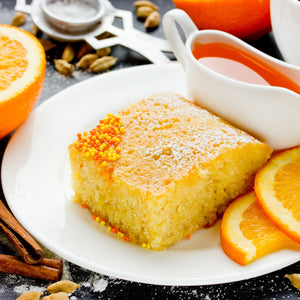 A slice of Palestinian Semolina & Fenugreek Cake with Orange Blossom on a white plate with fresh orange slices and a jug of sauce