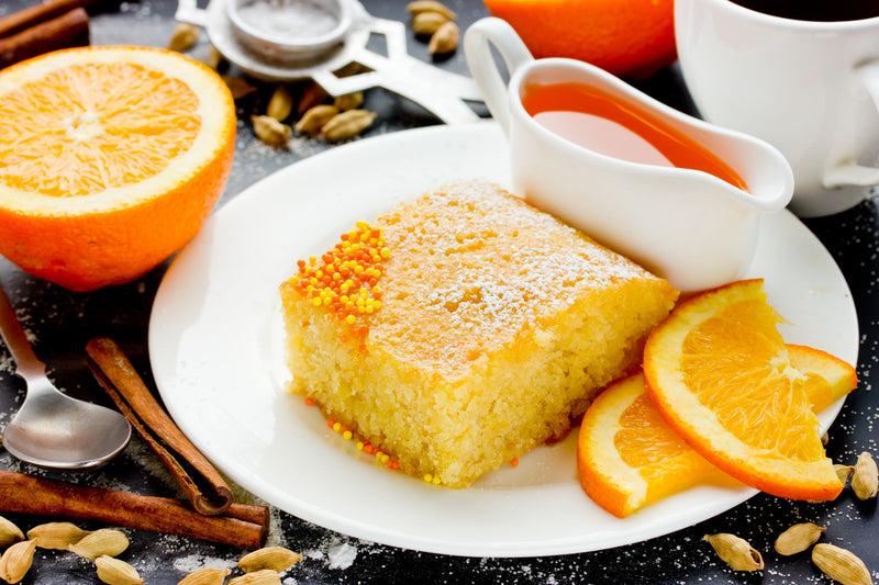 A slice of Palestinian Semolina & Fenugreek Cake with Orange Blossom on a white plate with fresh orange slices and a jug of sauce