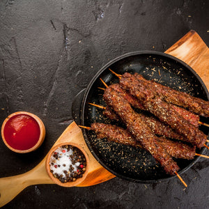 Harissa Lamb Kebabs on skewers on a sizzling plate with salt and black peppercorns and a pot of BBQ sauce. 