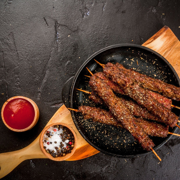 Harissa Lamb Kebabs on skewers on a sizzling plate with salt and black peppercorns and a pot of BBQ sauce. 