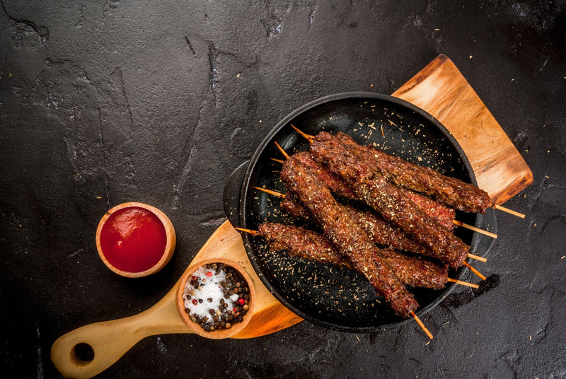 Harissa Lamb Kebabs on skewers on a sizzling plate with salt and black peppercorns and a pot of BBQ sauce. 