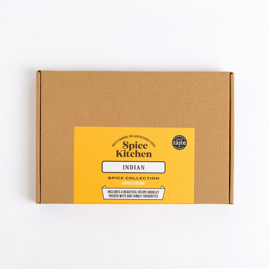 A square brown cardboard box for Spice Kitchen Indian Spice Collection, featuring a bright yellow label on the front with black text, photographed on a clean white background.
