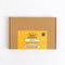 A square brown cardboard box for Spice Kitchen Indian Spice Collection, featuring a bright yellow label on the front with black text, photographed on a clean white background.