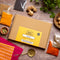 A flat-lay view of a 'Spice Kitchen' Indian Spice Collection box on a rustic wooden table, surrounded by various spices, ginger, cilantro, and kitchen tools.