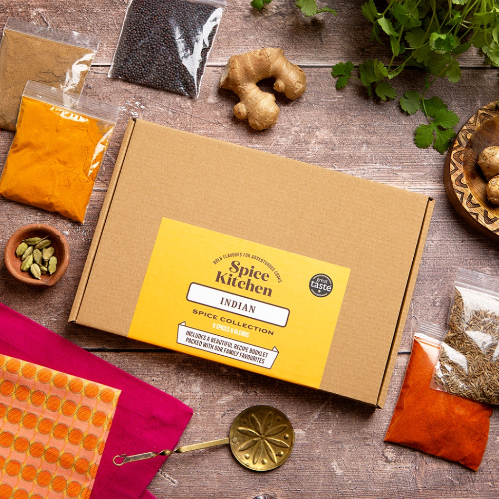 A flat-lay view of a 'Spice Kitchen' Indian Spice Collection box on a rustic wooden table, surrounded by various spices, ginger, cilantro, and kitchen tools.