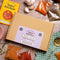 A flat-lay of a Spice Kitchen Middle Eastern Spice Collection kit, featuring a brown cardboard box with a lavender label, surrounded by various spice bags, garlic cloves, a recipe book, and a spoon on a rustic wooden table.