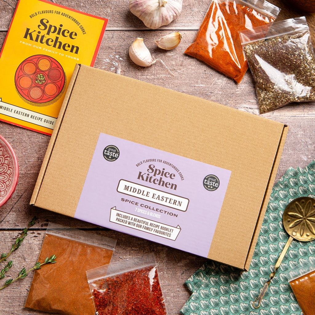 A flat-lay of a Spice Kitchen Middle Eastern Spice Collection kit, featuring a brown cardboard box with a lavender label, surrounded by various spice bags, garlic cloves, a recipe book, and a spoon on a rustic wooden table.