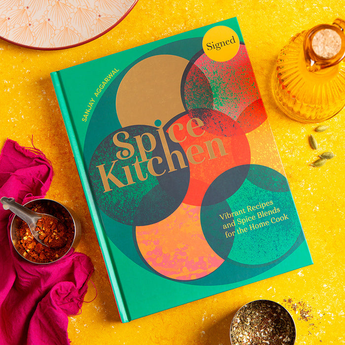 Spice Kitchen cookbook by Sanjay Aggarwal, on a yellow background with spices and a pink cloth.