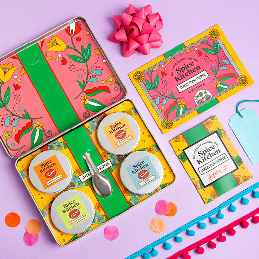 Colorful Spice Kitchen Feasting Kit product set with tins, packaging, recipe cards, a shopping list and a pink bow on a purple background.