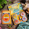 Open shot of Spice Kitchen's Feasting Tin – A Story of Spice' - showing four signature spice blends, a recipe booklet presented on a wooden chopping board with spices and silk sari material. 