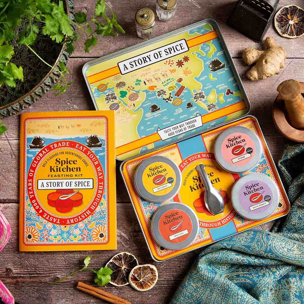 Open shot of Spice Kitchen's Feasting Tin – A Story of Spice' - showing four signature spice blends, a recipe booklet presented on a wooden chopping board with spices and silk sari material. 