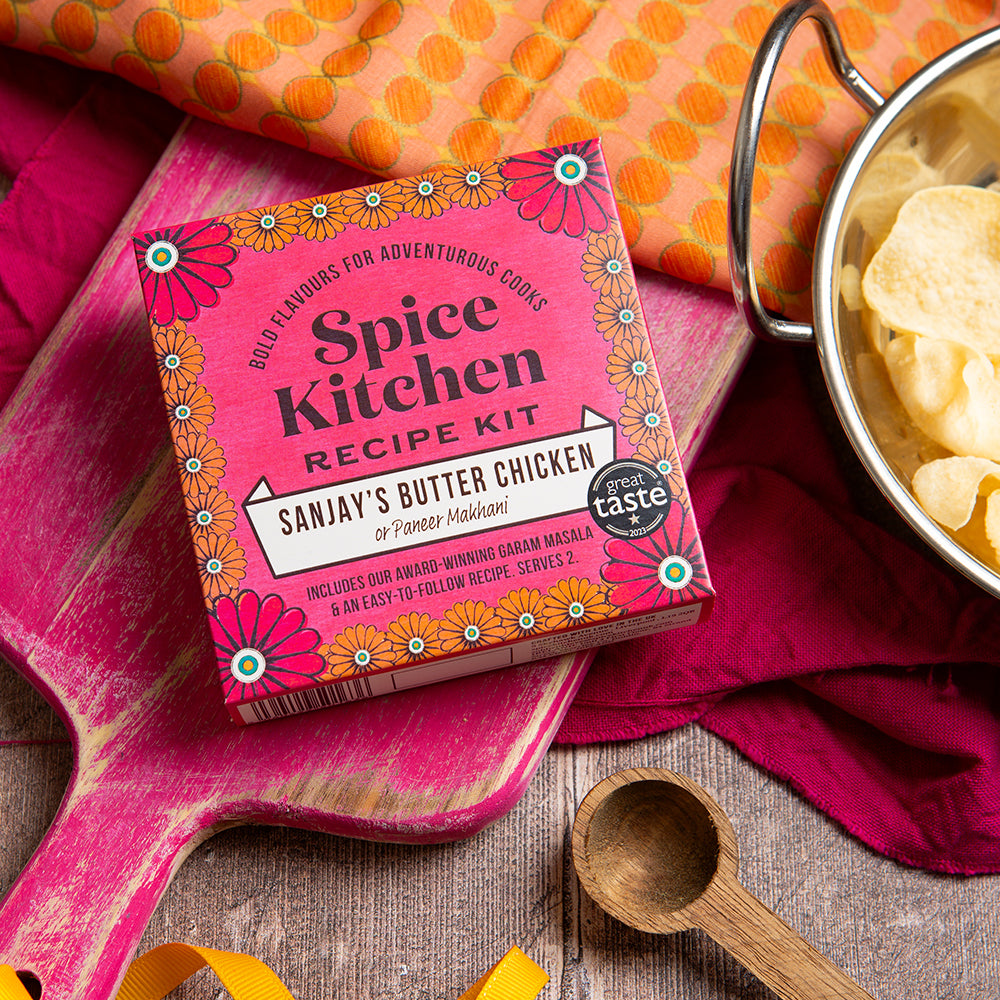 Spice Kitchen Recipe Kit for Sanjay's Butter Chicken on a pink cutting board with a pot of food and wooden spoon.