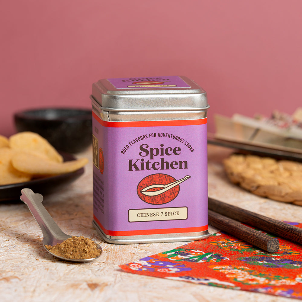 Spice Kitchen Chinese 7 Spice canister on a table with a spoonful of spice and snacks in the background.