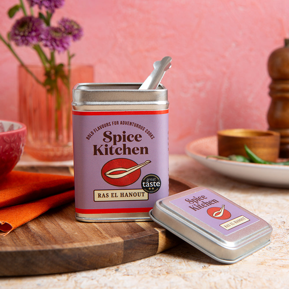 Spice Kitchen award winning Ras El Hanout signature spice blend tin with a spice measuring spoon on a kitchen counter with a pink background