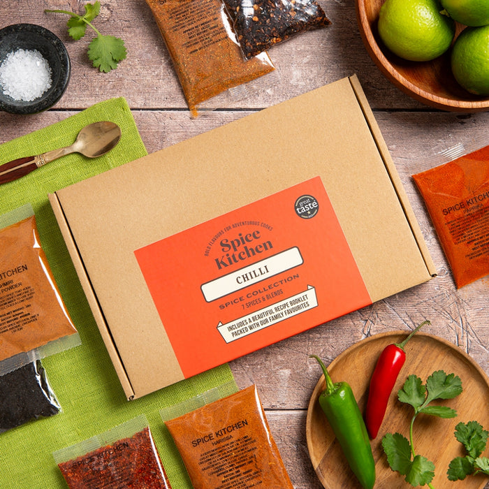 A Spice Kitchen Chilli Spice Collection box in kraft brown with a bright orange label, surrounded by loose spices, lime slices, jalapeños, cilantro, and seasoning bowls on a rustic wood surface.