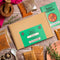 Spice Kitchen World spice collection gift box set arranged on a rustic wooden table with a kraft paper box, green Spice Kitchen branding, multiple spice pouches, a recipe guide booklet, dried orange slices, herbs, a spoon, and a wooden mortar and pestle.