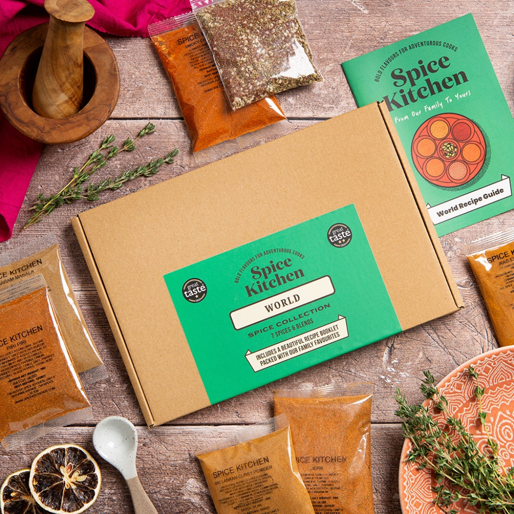Spice Kitchen World spice collection gift box set arranged on a rustic wooden table with a kraft paper box, green Spice Kitchen branding, multiple spice pouches, a recipe guide booklet, dried orange slices, herbs, a spoon, and a wooden mortar and pestle.