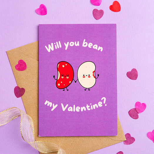 Purple Valentine's Day card with cartoon beans and 'Will you bean my Valentine?' text, surrounded by heart-shaped confetti on a purple background.