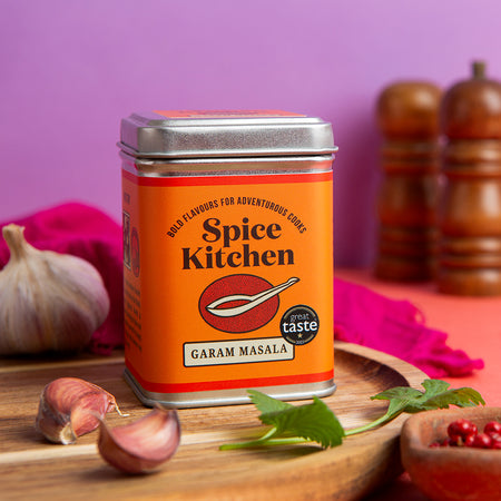 Spice Kitchen Garam Masala tin on a wooden board with garlic and herbs, against a purple background.