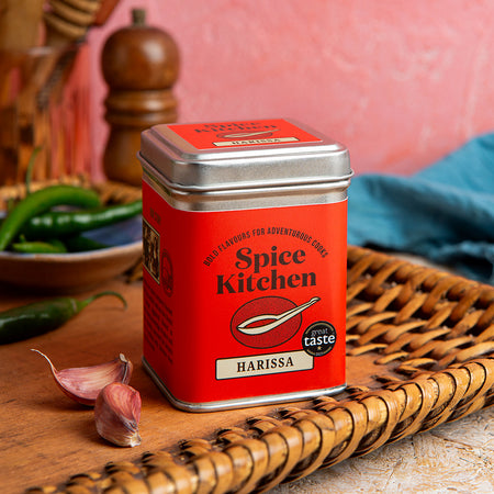 Spice Kitchen Harissa spice tin on a wooden surface with peppers and garlic in the background