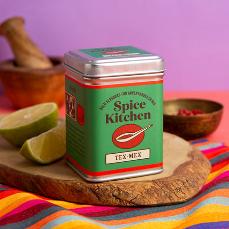Spice Kitchen Tex-Mex spice tin on a wooden board with limes and a mortar and pestle.