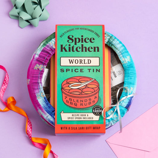 A gift set featuring a 'Spice Kitchen World Spice Tin' recipe book and spice blends, wrapped in a colorful patterned silk gift wrap and tied with a white ribbon, set against a lavender background with decorative ribbons and a bow.