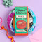 A gift set featuring a 'Spice Kitchen World Spice Tin' recipe book and spice blends, wrapped in a colorful patterned silk gift wrap and tied with a white ribbon, set against a lavender background with decorative ribbons and a bow.