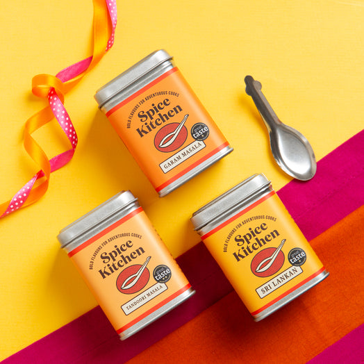 Three tins of Spice Kitchen's signature spice blends on a colourful background with a spice spoon including Sri Lankan Curry Powder, Garam Masala and Tandoori Masala – all Great Taste Award Winners