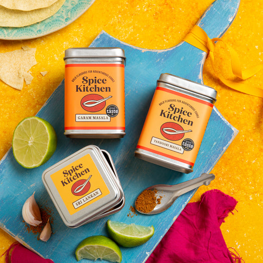 Three tins of 'Spice Kitchen' on a blue cutting board with lime, garlic, and tortilla chips on a yellow background.