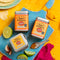 Three tins of 'Spice Kitchen' on a blue cutting board with lime, garlic, and tortilla chips on a yellow background.