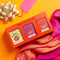 A gift set of three 'Chilli Collection' spice blends in a bright orange box, styled with a pink scarf, a gold bow, and a blank pink tag on a vibrant orange background.