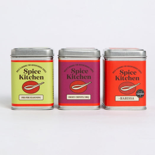 Three spice seasoning tins from Spice Kitchen lined up on a white background. The silver tins have colourful labels and include: Piri Pirir Seasoning, Smoky Chipotle BBQ, and Harissa.