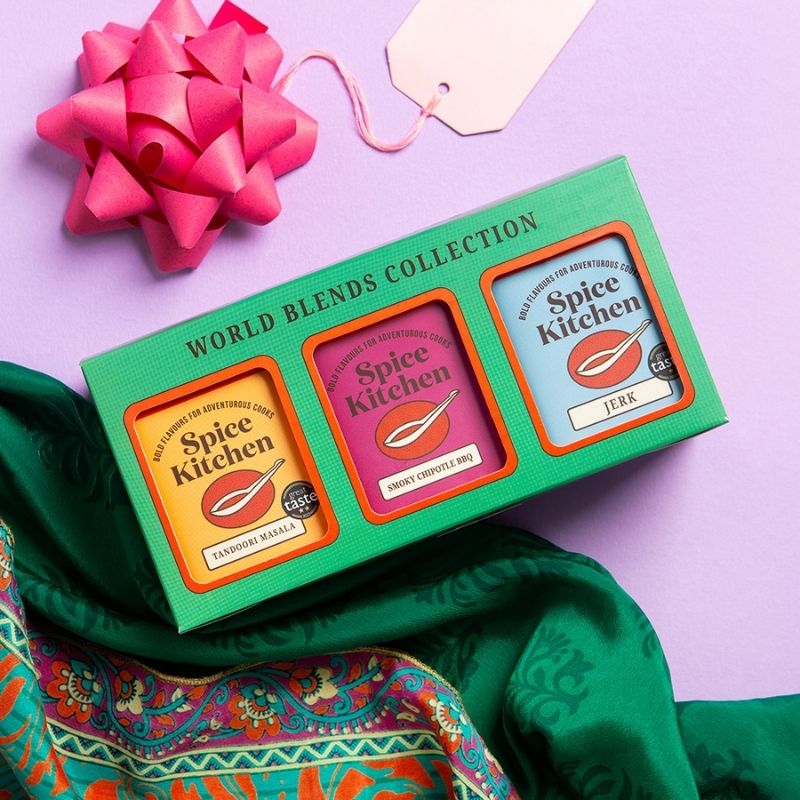 A green gift box labeled 'WORLD BLENDS COLLECTION' containing three Spice Kitchen seasoning tins, styled on a lavender background with a pink bow, cream tag, and green patterned fabric.